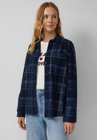 Marineblauwe geblokte blouse met knoopsluiting, lange mouwen en zakken. Draagt over een lichtgekleurde T-shirt en gecombineerd met lichte jeans.