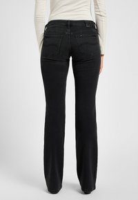 Frau in schwarzen geraden Jeans und einem weißen langärmligen Oberteil, von hinten vor einem schlichten Hintergrund gezeigt.