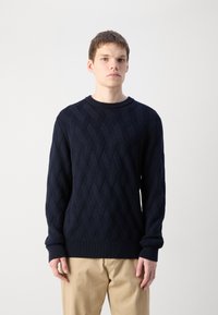 Il maglione lavorato a maglia blu navy presenta un motivo a rombi testurizzato, collo a giro e polsini a coste. Abbinato a pantaloni beige chiaro.