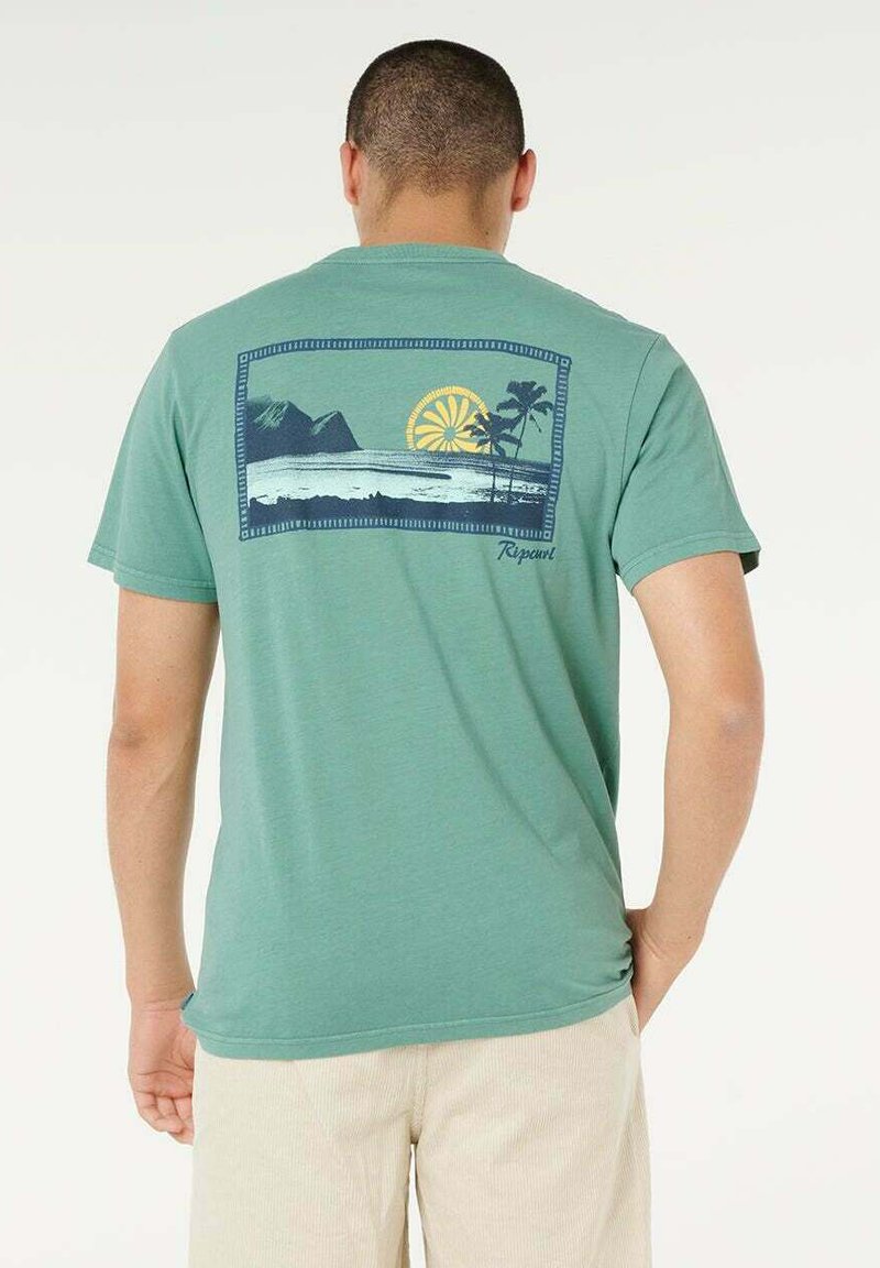Uomo che indossa una t-shirt verde con una grafica sul retro raffigurante oceano, sole, palme e montagne, abbinata a pantaloni beige chiaro.