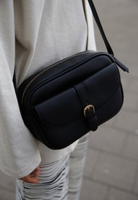 Bolso bandolera de cuero negro con acabado texturizado, herrajes dorados y detalle de hebilla en la parte frontal. Cierre de cremallera en la parte superior.