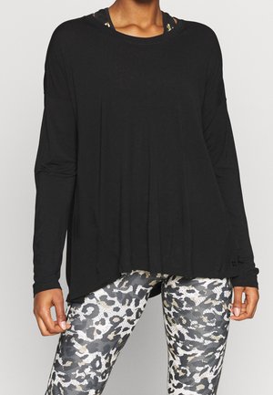 Persoon die een losse zwarte top met lange mouwen draagt over zwart-witte gebloemde leggings, staand tegen een effen lichte achtergrond.