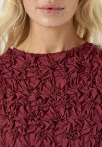 Mørkerød topp med teksturert stoff som har rynket og bølgete detaljer over hele, med rund hals som fremhever det unike overflatedesignet.