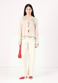 Blouse beige clair avec broderie florale verte et finition volants, associée à un pantalon large crème et des escarpins rouges, tenant un sac rouge.