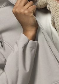 Ruka držící límec světle šedé bundy s fleecovou podšívkou, částečně rozepnuté, zblízka ukazuje detaily rukávu a zipu.