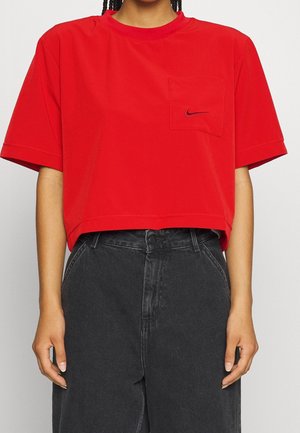 Persona che indossa una maglietta rossa corta con un piccolo logo Nike sulla tasca del petto e jeans neri a vita alta.