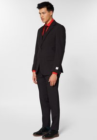 Completo nero con giacca aderente, camicia rossa e scarpe eleganti nere. Dettagli notevoli includono stringhe blu e un piccolo logo sulla manica.