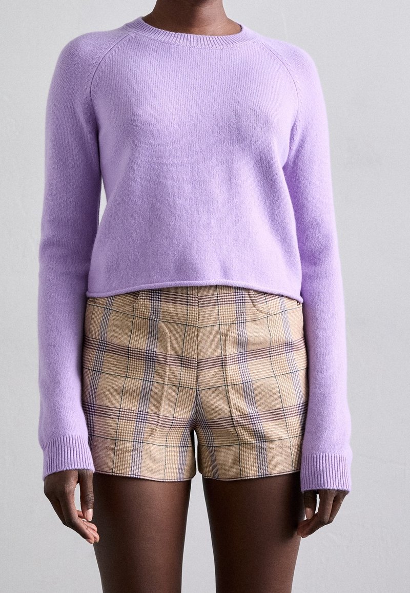 Personne portant un pull à manches longues violet pastel et un short taille haute à carreaux beige sur un fond uni.