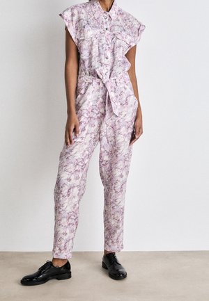 Kvinde iført en ærmeløs jumpsuit med pink og hvidt mønster med et bundet bælte i taljen og sorte snørestøvler, der står på en neutral gulv.