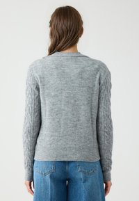 Grijze gebreide trui met een geribbelde kraag en kabelbreipatroon op de mouwen. Zachte textuur, slanke pasvorm bij de taille, gecombineerd met hoge taille jeans.