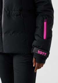 Giacca a vento nera con una tasca con cerniera rosa e una fascia con logo sulla manica. Texture liscia con un design aderente.