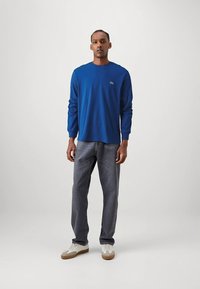 Kék hosszú ujjú ing, kerek nyakkivágással és zöld logóval a mellkasnál, szürke farmerrel és bézs részletekkel ellátott fehér tornacipővel párosítva.