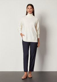 Cremfarbener Rollkragenpullover mit gerippten Bündchen, kombiniert mit dunkelblauen Hosen und hellbraunen Slippern mit Kettenakzent. Einfaches und dezentes Farbschema.
