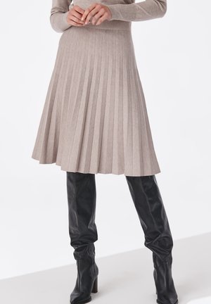 Femme portant une jupe mi-longue plissée beige et des bottes en cuir noires à talons montant jusqu'aux genoux, les mains jointes à la taille.