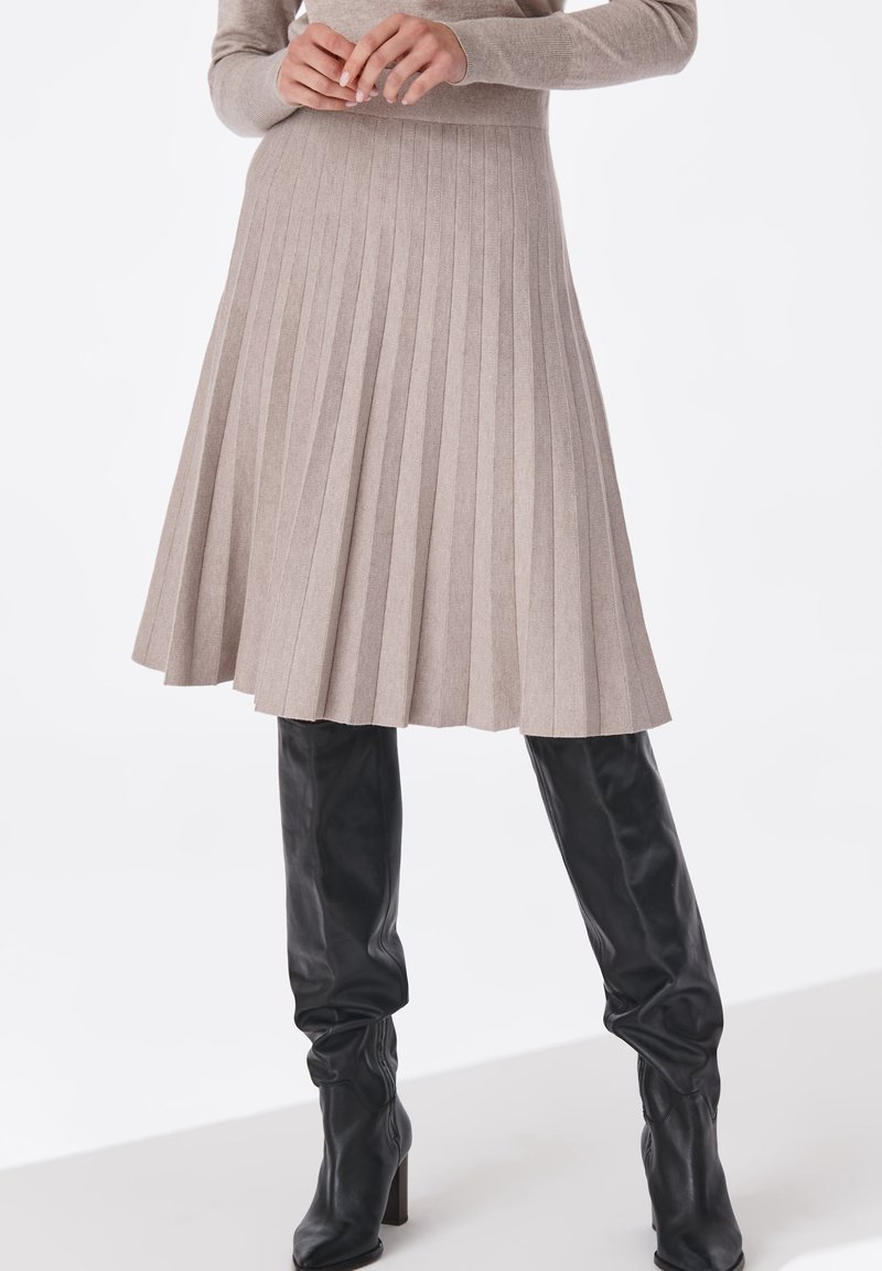 Femme portant une jupe mi-longue plissée beige et des bottes en cuir noires à talons montant jusqu'aux genoux, les mains jointes à la taille.