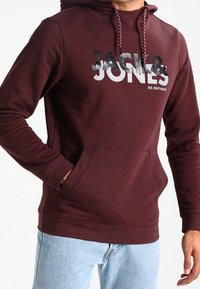 Felpa bordeaux con tasca frontale, cappuccio con lacci e stampa grafica "JACK & JONES RE:DEFINED" in colori a contrasto. Tessuto misto cotone.