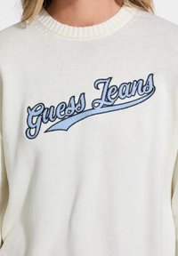 Pulover alb tricotat, cu inscripția "Guess Jeans" brodată în culori bleumarin și albastru deschis pe piept. Guler și mansete cu nervuri.