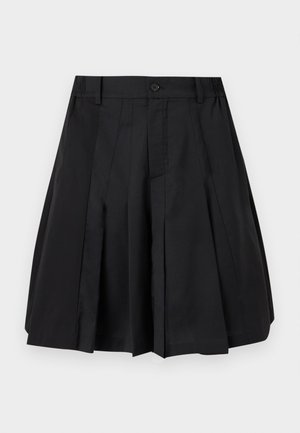 Homme portant une casquette bleue, une veste marron et noire, un polo gris, une jupe plissée noire, des chaussettes noires, des chaussons beige et portant un petit sac noir.