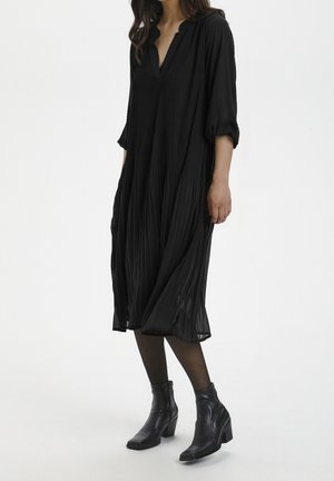 Femme portant une robe midi plissée noire avec manches retroussées, des collants noirs et des bottines noires à talons, sur un fond blanc uni.