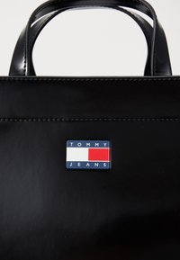 Schwarze glänzende Tragetasche mit doppelten Griffen und einem rechteckigen Tommy Jeans Logo-Abzeichen in Rot, Weiß und Blau.