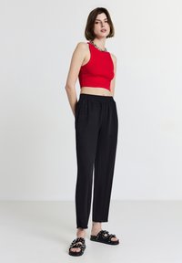 Femme portant un crop top rouge sans manches, un pantalon noir taille haute, des sandales noires à clous, se tenant contre un fond blanc.