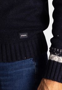 Marineblauwe gebreide trui met geribbelde onderkant en manchetten. Bevat een witte en grijze geaccentueerde patroon op de mouwen. Inclusief een merketiket.