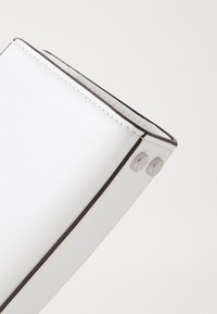 Valkoinen nahkalaukku kulmassa, jossa näkyy ompeleet ja kaksi hopeista metallikynnen päätä, yksinkertaisella valkoisella taustalla.