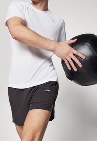 Vitt kortärmat träningsskjorta med ett strukturerat mönster, parat med svarta shorts med sidorek och en diskret logotyp, håller en svart medicinboll.