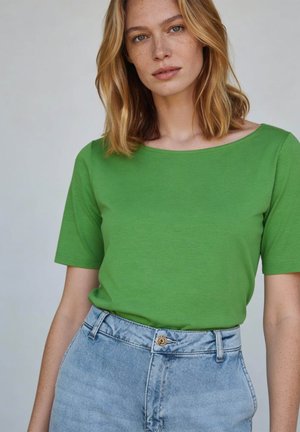 Jonge vrouw met schouderlang blond haar, een groene top met korte mouwen die is ingestopt in een lichtblauwe spijkerbroek met hoge taille.