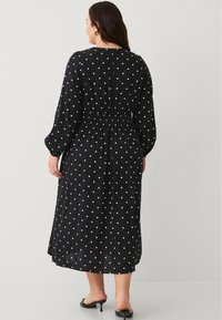 Schwarzes Kleid mit weißen Polka-Punkten, langen Ärmeln, gerafftem Taillenbund und einer Länge bis zur Mitte der Waden. Der Stoff scheint leicht und hat eine glatte Textur.