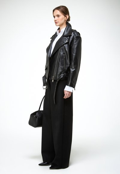 Mlada žena u crnoj kožnoj jakni preko bijele majice, u crnim hlačama širokih nogavica, držeći crnu torbicu, stoji bočno.