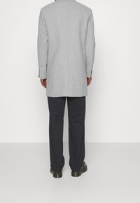 Mężczyzna stojący tyłem, ubrany w jasnoszary płaszcz, ciemne spodnie i czarne buty na jednolitym białym tle.