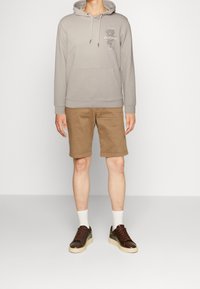 Sudadera con capucha de color gris claro, con un bolsillo tipo canguro, combinada con pantalones cortos color arena. Zapatillas marrones con suelas beige. Calcetines blancos visibles.