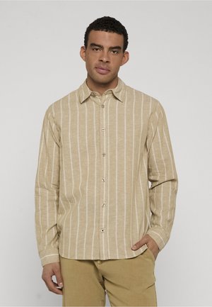 Homme portant une chemise beige à boutons avec des rayures verticales blanches et un pantalon marron clair, debout avec une main dans la poche devant un fond uni.