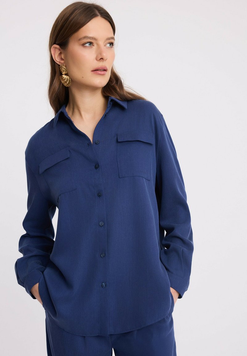 Kobieta w niebieskiej koszuli na guziki z kieszeniami z przodu i dopasowanych spodniach, z rękami w kieszeniach, patrząca w bok na tle jednolitego tła.