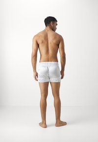 White swim shorts made of lightweight fabric with elastic waistband, seen from the back on a person with bare feet and no shirt.