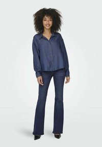 Camicia button-up in denim scuro con colletto e maniche lunghe, abbinata a jeans scuri a zampa d'elefante e tacchi neri; texture liscia, vestibilità rilassata.