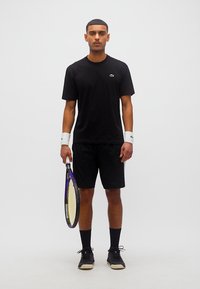 Άνδρας με μαύρη αθλητική ενδυμασία που κρατά ρακέτα τένις, φοράει λευκά περιβραχιόνια και μαύρα αθλητικά παπούτσια, στέκεται μπροστά σε απλό φόντο.