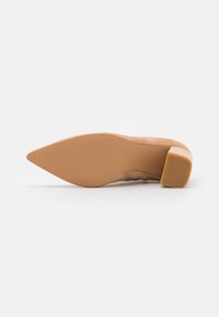 Scarpa alla moda beige in suede con tacco alto, punta affusolata e tacco spesso, che mette in evidenza una superficie liscia e textured e una suola curvata.