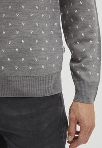 Un pull en tricot gris présentant un motif blanc répétitif. Poignets et ourlet côtelés, avec une étiquette sur le côté. Porté sur un pantalon sombre.