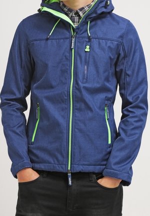 Mand iført en blå hættetrøje med neon grønne lynlåse og snore, hænderne i lommerne, og en ternet skjorte under.