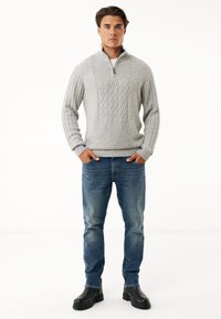 Grau gestrickter Zip-Pullover mit Kabelmuster auf der Vorderseite, kombiniert mit blauen Jeans und schwarzen Stiefeln, vor einem weißen Hintergrund stehend.