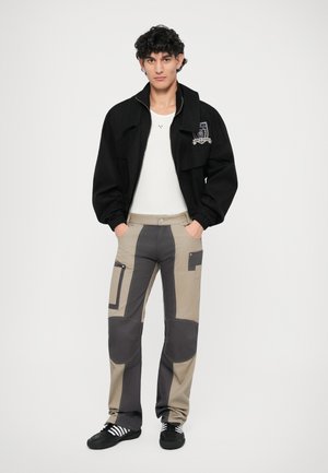 Giacca nera con zip e patch, camicia bianca aderente, pantaloni beige con pannelli grigi e tasche, scarpe nere con strisce bianche.