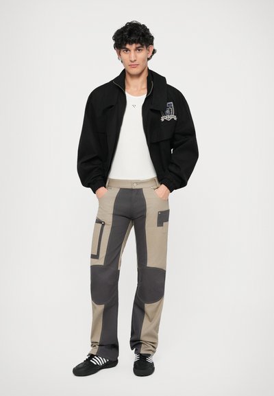 Zwarte zip-up jacket met patch, getailleerd wit shirt, beige broek met grijze panelen en zakken, zwarte schoenen met witte strepen.