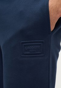 Κοντινή λήψη υφάσματος σε ναυτικό μπλε με ανάγλυφο λογότυπο Lacoste Paris κοντά στο χέρι ενός ατόμου και στη ραφή της πλευράς.