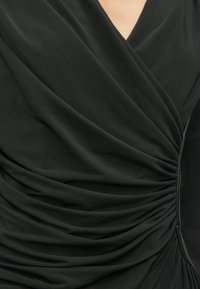 Melnas auduma blūze ar mīkstiem, krītieniem un ručinga detaļām. Ar dziļu V veida kakla izgriezumu un gludu tekstūru.