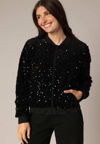 Veste bomber noire en tissu texturé et duveteux avec des embellissements en sequins. Elle présente une fermeture éclair à l'avant et des poignets et un ourlet côtelés.