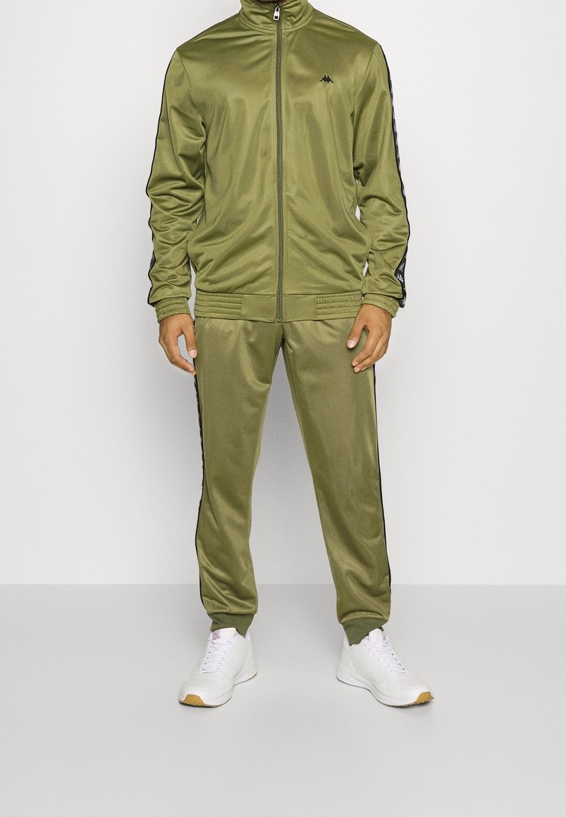 Homme portant une veste de survêtement zippée vert olive et un pantalon assorti avec des bandes latérales noires et des baskets blanches, debout devant un fond blanc.