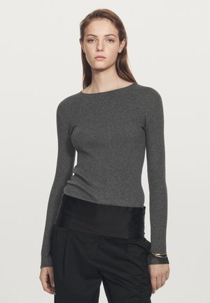 Ung kvinde med langt hår iført en tætsiddende grå ribstrikket sweater og sorte bukser, stående foran en ensfarvet lys baggrund.