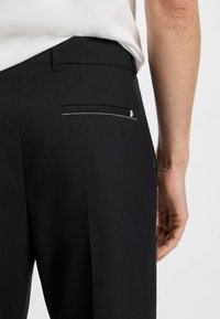 Black tailored trousers with a smooth texture, featuring a single back pocket with contrasting white stitching and a metallic accent.